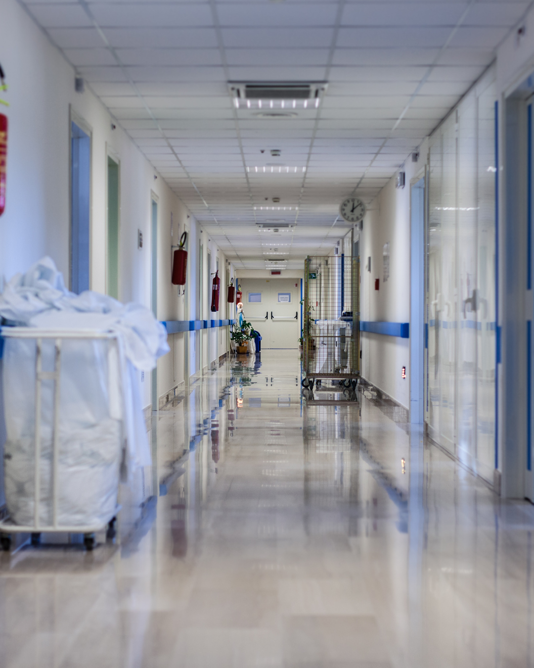 A shot of an empty hospital hallway with white walls and a few linen carts and fire extinguishers dotted along the edges of the hallway.